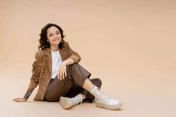 Donna sorridente in giacca scamosciata e pantaloni e stivali alla moda seduta su sfondo beige — Foto stock