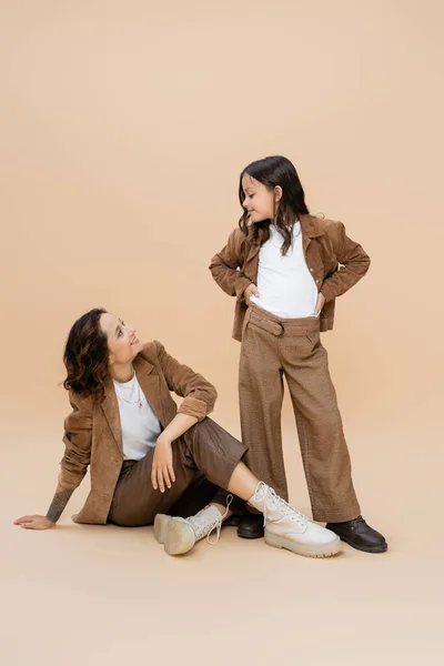 Happy woman in autumn clothes looking at trendy daughter posing with hands on hips on beige background — Stock Photo