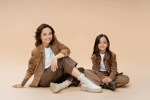 Madre e hija en traje de otoño de moda sentado sobre fondo beige - foto de stock