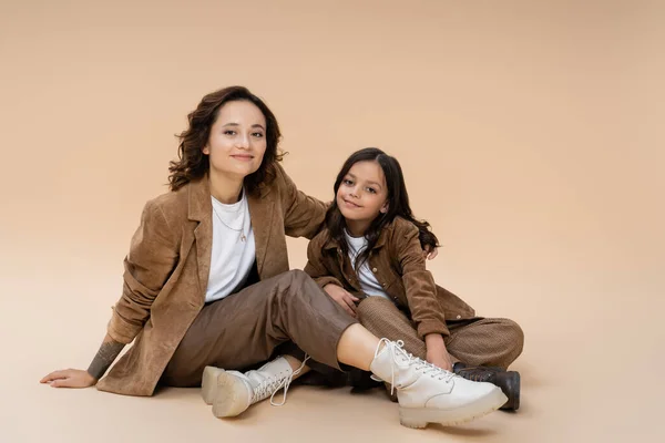 Mujer feliz en chaqueta marrón y botas de moda abrazando hija mientras está sentado sobre fondo beige - foto de stock