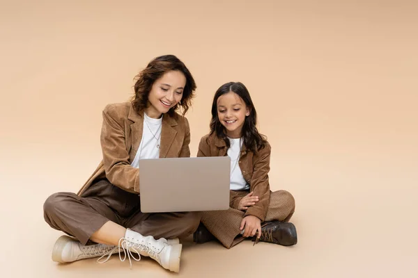 Smiling girl with mom in stylish autumn clothes sitting with crossed legs near laptop on beige background — Stock Photo