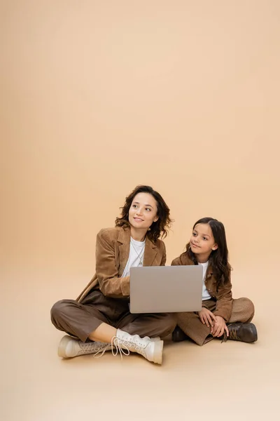 Femme souriante et élégante regardant loin près de l'ordinateur portable et la fille sur fond beige — Photo de stock