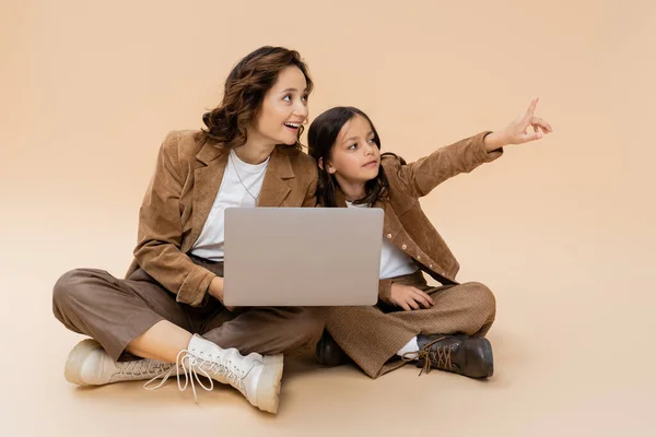 Mädchen in trendiger Herbstkleidung zeigt mit dem Finger auf Laptop und lächelnde Mutter auf beigem Hintergrund — Stockfoto