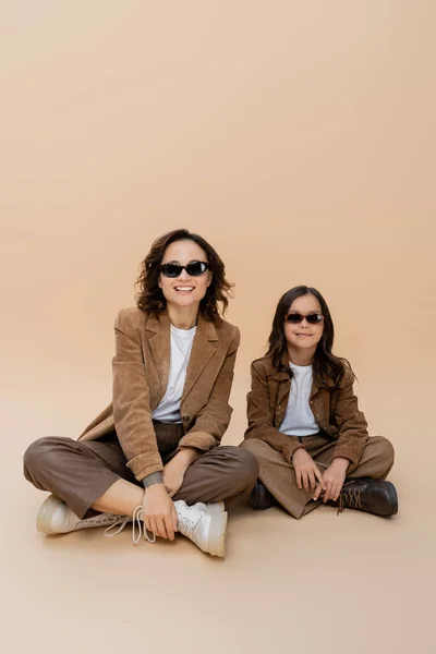 Fröhliches Mädchen und Mutter im stylischen Herbst-Outfit und Sonnenbrille sitzend mit überkreuzten Beinen auf beigem Hintergrund — Stockfoto