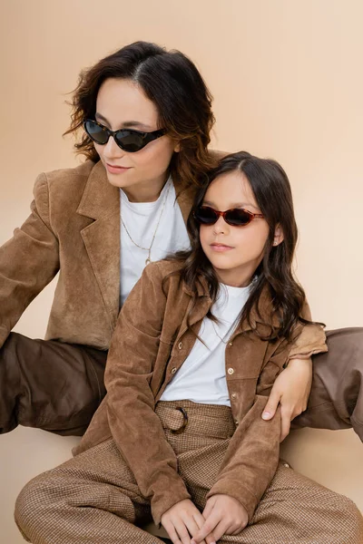 Madre e hija de moda en gafas de sol y chaquetas de gamuza marrón sentadas sobre fondo beige - foto de stock