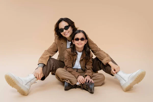 Fashionable mom and daughter in sunglasses posing in suede jackets and boots on beige background — Stock Photo