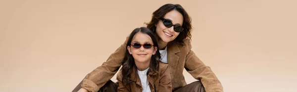 Mujer alegre y chica en gafas de sol y chaquetas de gamuza marrón aisladas en beige, bandera - foto de stock