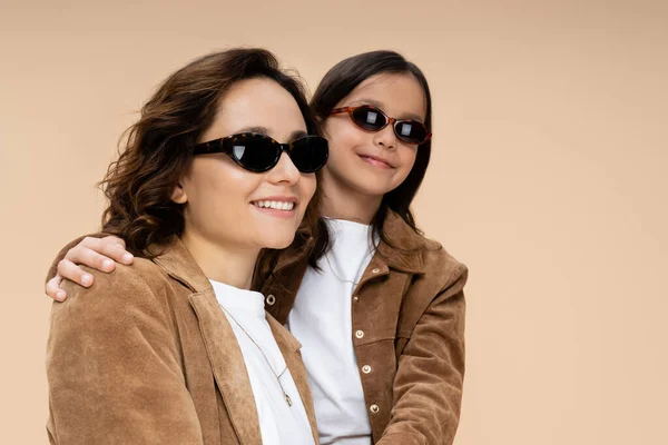 Madre e hijo en chaquetas de gamuza y gafas de sol elegantes sonriendo aislados en beige - foto de stock