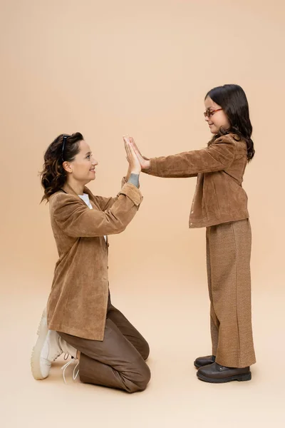 Side view of mother and daughter in stylish autumn outfit playing patty cake game on beige background — Stock Photo