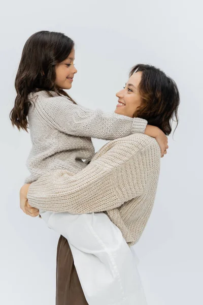 Vista lateral de la mujer sonriente en ropa de abrigo sosteniendo a la hija en manos aisladas en gris - foto de stock