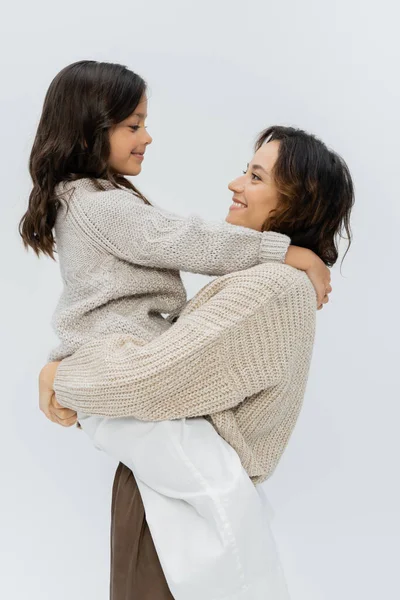 Vue latérale de la femme souriante en vêtements chauds tenant fille dans les mains isolées sur gris — Photo de stock