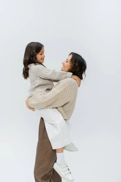 Vista lateral da mulher morena em roupas de outono elegantes segurando filha feliz isolada no cinza — Fotografia de Stock