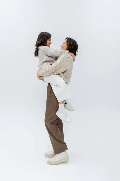 Vista lateral da mulher em roupas de outono elegantes segurando a filha em mãos no fundo cinza — Fotografia de Stock