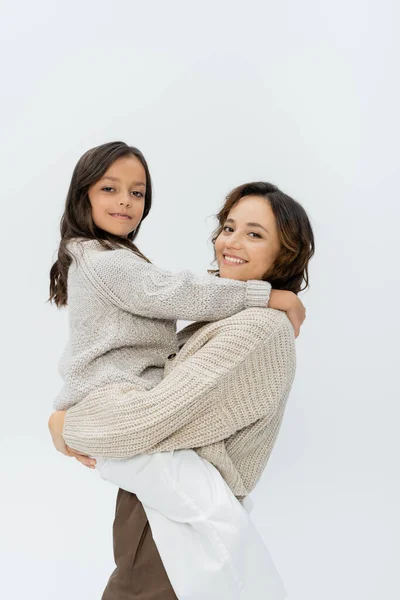 Mujer y niña sonrientes en prendas de punto abrazando y sonriendo a la cámara aislada en gris - foto de stock