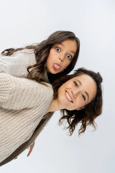 Sonriente mujer en punto piggybacking hija sobresaliendo lengua aislada en gris - foto de stock