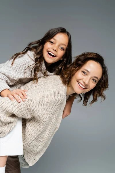 Happy brunette woman in knitwear piggybacking cheerful daughter isolated on grey — Stock Photo