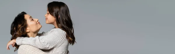 Vista lateral de la joven mujer haciendo pucheros labios mientras abraza hija feliz aislado en gris, bandera - foto de stock