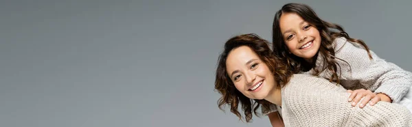 Brunette woman smiling at camera and piggybacking daughter isolated on grey, banner — Stock Photo