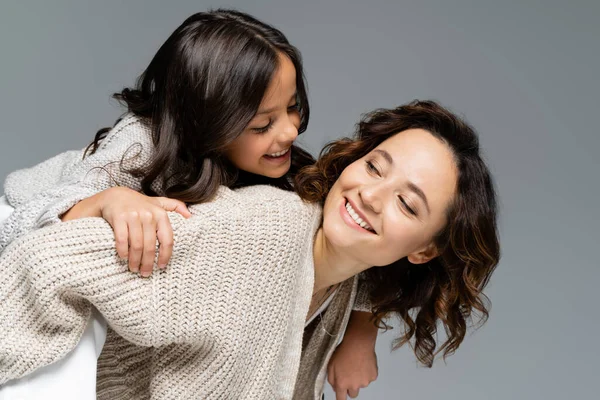Smiling woman in autumn clothes looking at happy daughter while piggybacking her isolated on grey — Stock Photo