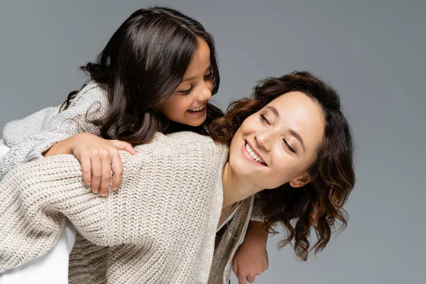 Sonriente mujer en otoño ropa mirando feliz hija mientras piggybacking su aislado en gris - foto de stock