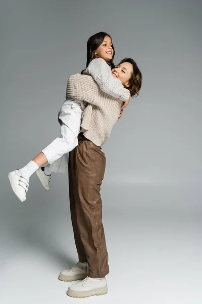 Vista lateral de la mujer a la moda sosteniendo a la hija alegre en las manos en gris - foto de stock