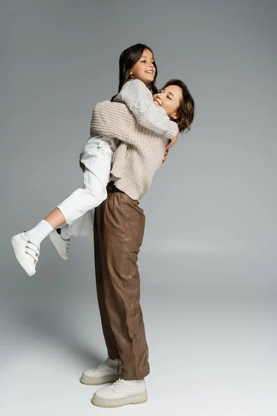 Vue latérale de la femme à la mode tenant fille gaie dans les mains sur gris — Photo de stock