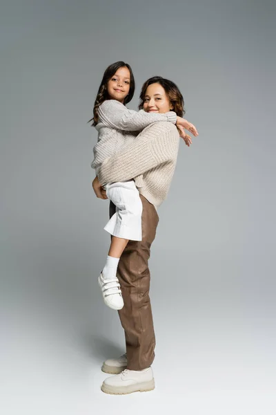 Happy and trendy woman holding smiling daughter in hands while looking at camera on grey background — Stock Photo