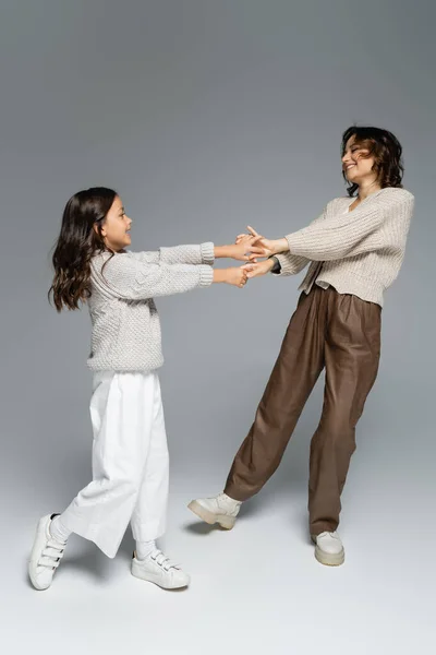 Excited woman with daughter in autumn outfit dancing while holding hands on grey background — Stock Photo
