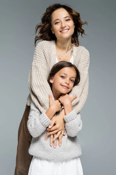 Eccitata donna bruna in maglieria alla moda abbracciando sorridente figlia isolata sul grigio — Foto stock