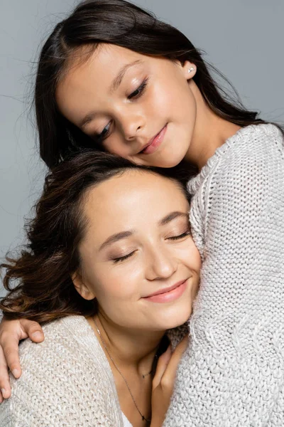 Morena chica abrazando madre sonriendo con los ojos cerrados aislado en gris - foto de stock
