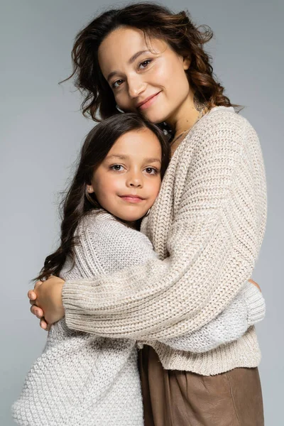 Cheerful mom and daughter in cozy knitwear hugging and smiling at camera isolated on grey — Stock Photo