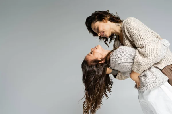 Vista lateral de la sonriente mujer tatuada abrazando a su hija mientras baila aislada en gris - foto de stock