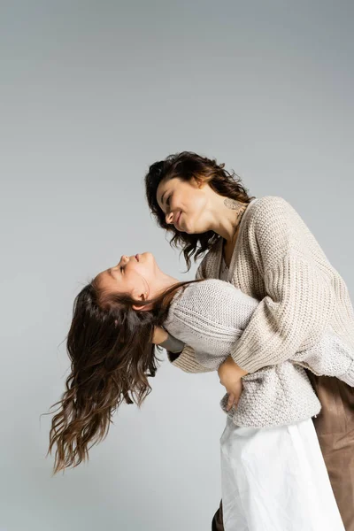 Seitenansicht einer lächelnden und stilvollen Frau, die ihre Tochter umarmt, während sie isoliert auf grau tanzt — Stockfoto