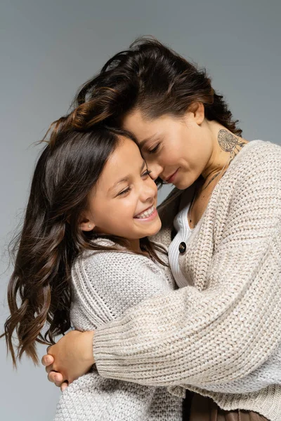 Tattooed woman in warm clothes embracing excited daughter isolated on grey — Stock Photo