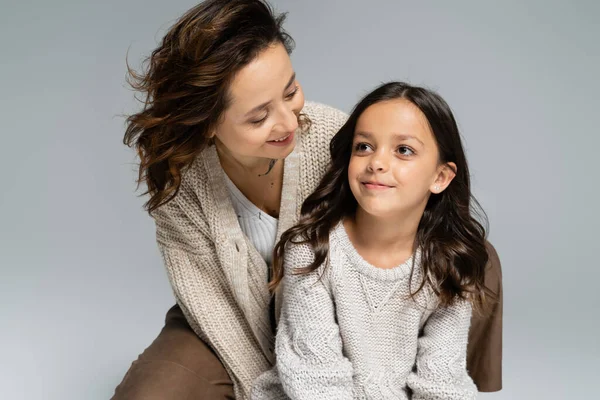 Cheerful brunette woman in trendy and warm clothes looking at smiling daughter isolated on grey — Stock Photo