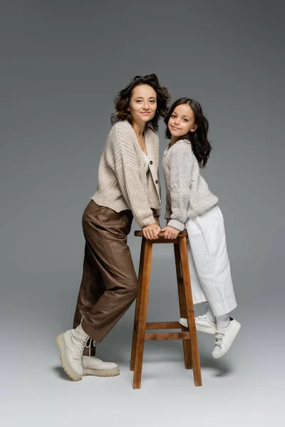 Longitud completa de la madre y la hija en traje de otoño de moda posando cerca de taburete de madera en gris - foto de stock