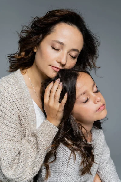 Brunette woman with closed eyes embracing head of daughter isolated on grey — Stock Photo