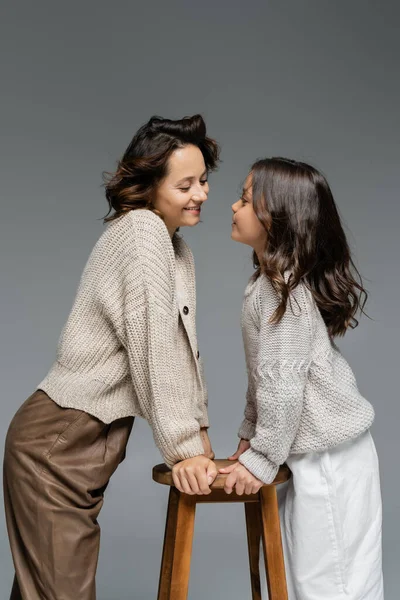 Madre e hija de moda en prendas de punto cálidas mirándose cerca de taburetes de madera aislados en gris - foto de stock