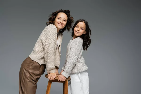 Mère et fille souriantes en tenue d'automne tendance posant près de tabouret en bois isolé sur gris — Photo de stock