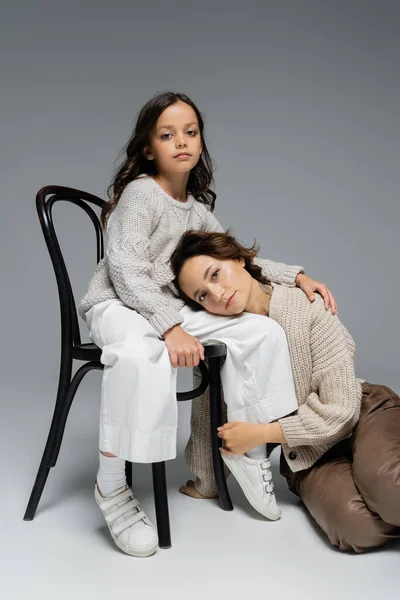 Stylish woman leaning on daughter sitting on chair and looking at camera on grey background — Stock Photo