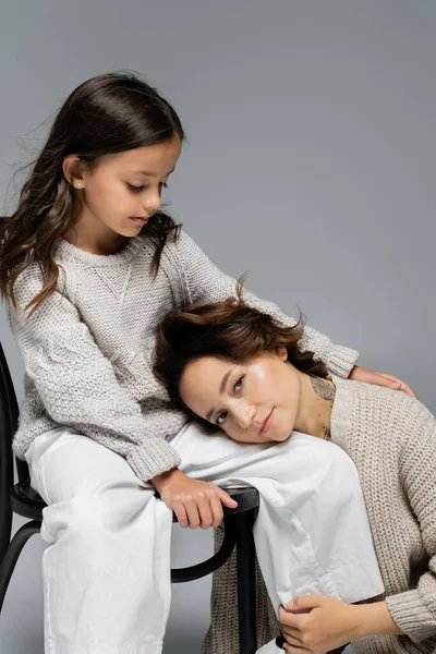 Brünette Frau blickt in die Kamera, während sie sich auf trendige Tochter stützt, die auf Stuhl auf grauem Hintergrund sitzt — Stockfoto