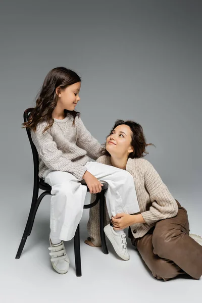 Mulher feliz olhando para a filha sentada na cadeira na roupa de outono na moda em fundo cinza — Fotografia de Stock