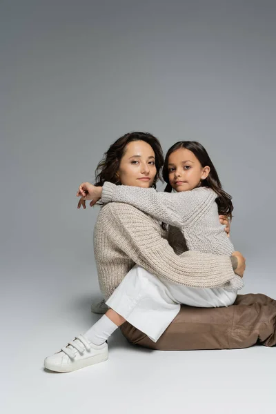 Женщина и ребенок в теплых трикотажах обнимаются и смотрят в камеру на сером фоне — стоковое фото
