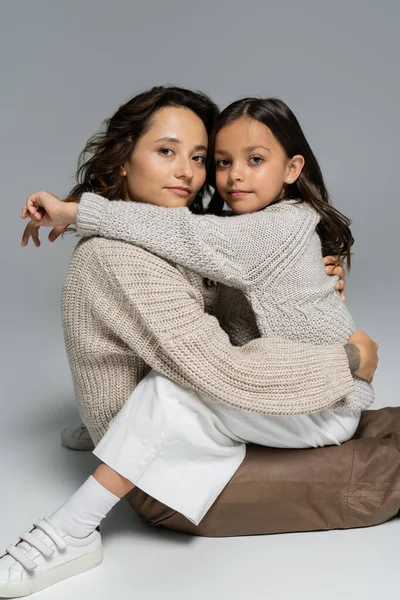 Мати і дочка в теплому одязі обіймаються і дивляться на камеру на сірому фоні — стокове фото