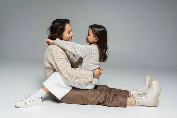 Seitenansicht der brünetten Mutter und Tochter in warmer Kleidung, die einander auf grauem Hintergrund anlächeln — Stockfoto