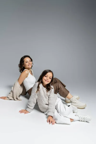 Mother and daughter in fashionable autumn outfit smiling at camera while sitting on grey background — Stock Photo