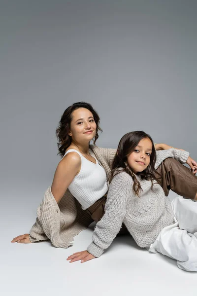 Brunette woman and girl in knitwear smiling at camera while sitting on grey background — Stock Photo