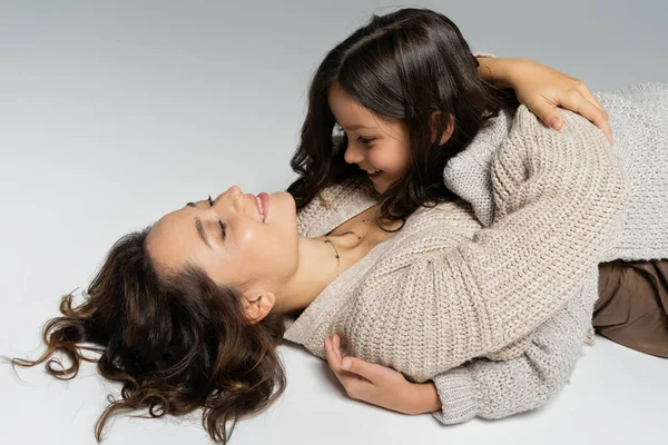 Sonriente mujer en cálido cárdigan abrazando hija mientras está acostado sobre fondo gris - foto de stock