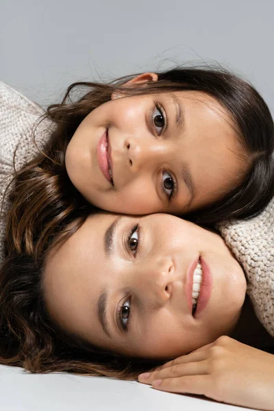 Feliz madre e hija en prendas de punto mirando a la cámara mientras yacen aisladas en gris - foto de stock
