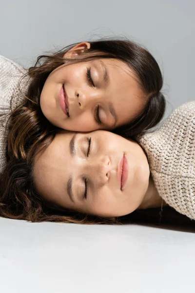 Madre e hija en suéteres cálidos acostadas con los ojos cerrados sobre fondo gris - foto de stock